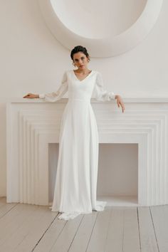 a woman standing in front of a fireplace wearing a white dress with long sleeves and high slit