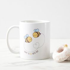 a white coffee mug with two bees on it next to a donut that says happy to be with you