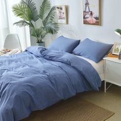 a bed with blue sheets and pillows in a white room next to a potted plant