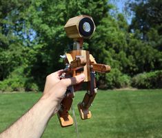 a hand holding a wooden toy robot in front of some green grass with trees in the background