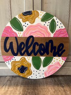 a wooden sign that says welcome with flowers and leaves painted on the front, sitting against a wood background