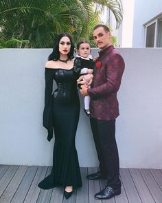 the man and woman are posing with their baby in front of a white wall outside