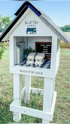 a white birdhouse with eggs for sale on the front and side in an open field