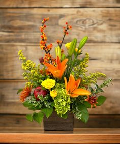 an arrangement of flowers and greenery in a square vase on a wooden table against a wood wall