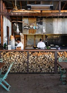 two men sitting at a bar with stacks of firewood behind them in a restaurant