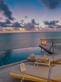 two lounge chairs sitting on top of a wooden deck next to the ocean at sunset