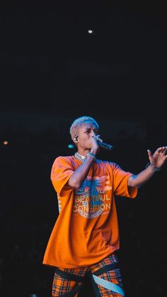 a male in an orange shirt and plaid pants is singing into a microphone on stage