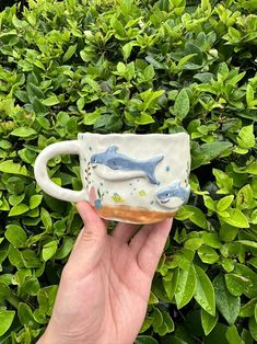 a hand holding a coffee cup with two dolphins on it in front of some bushes