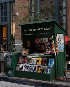 a green kiosk selling magazines on the side of a street in front of a brick building