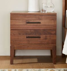 a wooden nightstand with two drawers and a white vase sitting on top of it next to a bed