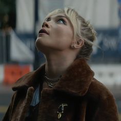 a woman with blonde hair wearing a brown fur coat looking up into the sky while standing on a city street