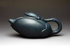 a teapot shaped like a fish on a gray surface with one eye open and the other half closed