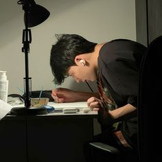 a young man writing on a piece of paper in front of a desk with a lamp