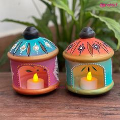 two colorful candle holders sitting next to each other on top of a wooden table in front of a potted plant