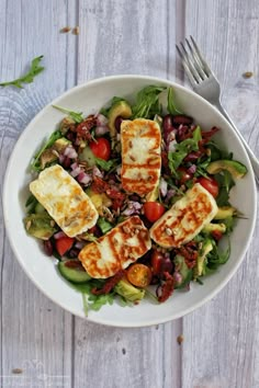 a white bowl filled with salad and tofu on top of a wooden table next to a fork