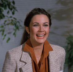 a smiling woman in a blazer and orange shirt smiles at the camera while holding a cup