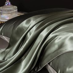 a bed covered in a green satin comforter next to a night stand with a glass jar on it