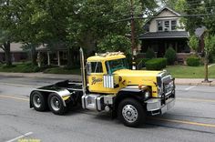 a large yellow truck driving down a street next to a tall house and trees on the side of the road