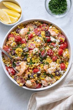 a bowl filled with shrimp, corn and tomatoes next to lemon wedges on a white surface