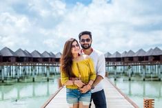 a man and woman standing on a dock near the water with huts in the background