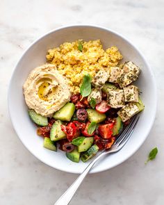 a white bowl filled with vegetables and hummus next to a fork on a marble surface