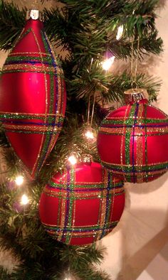 two red ornaments hanging from a christmas tree with lights on it's branches and one has a plaid pattern