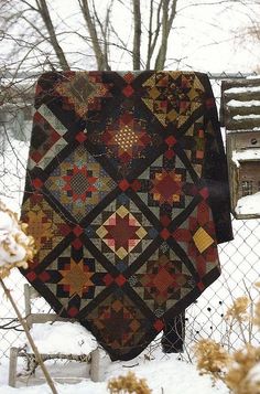 a quilt is hanging on a fence in the snow, with some flowers behind it