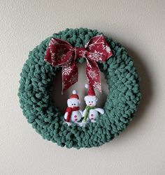 a green wreath with two snowmen and a red bow hanging on the wall next to a white wall