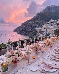 a table set up with flowers and candles for an outdoor dinner overlooking the ocean at sunset