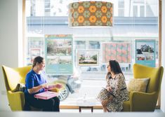 a man and woman sitting in chairs talking to each other near a window with pictures on it