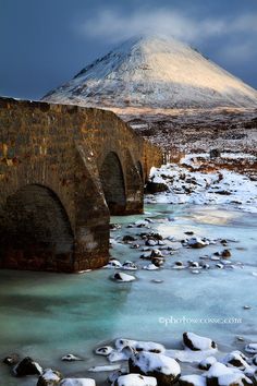 an old stone bridge over a river with snow on the ground and a mountain in the background