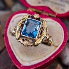 This lovely vintage ring features a lab created blue spinel in a full bezel setting that is decorated with a foliate motif halo. The ring is crafted in patinated 10k yellow gold and is currently a size 6.5. Vintage Blue Sapphire Ring With Bezel Setting, Vintage Blue Sapphire Ring With Halo Setting, Vintage Blue Ring With Bezel Setting, Vintage Blue Cluster Ring With Center Stone, Antique Gold Topaz Ring With Center Stone, Blue Vintage Cluster Ring With Center Stone, Vintage Blue Cluster Ring For Formal Occasions, Vintage Blue Gemstone Cluster Ring, Blue Gemstone Cluster Ring Vintage Style