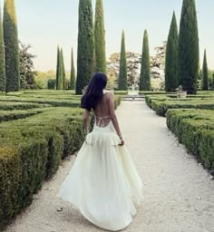 a woman in a white dress walking down a path lined with hedges and trees,