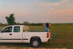 a woman standing on the back of a white truck in a grassy field next to a road