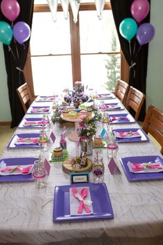 a table set up with purple and pink plates, napkins, and balloons in the background