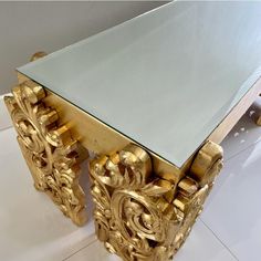 an ornately designed table with glass top and gold leaf decorations on the legs, against a white tiled floor