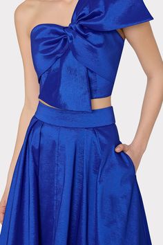 Cut from a bright blue, luxurious and crisp taffeta fabric, this voluminously full skirt falls to a modest, midi length. It has two side pockets and a zipper in the back to close. For the full look, pair it with its coordinated Laney Bow Crop Top. Party Satin Skirt With Folds, Blue Satin Skirt For Spring, Elegant Cropped Flowy Skirt, Blue Satin Evening Skirt, Elegant Blue Skirt For Prom, Blue Gathered Skirt For Party, Blue Voluminous Skirt For Parties, Voluminous Blue Skirt For Party, Bow Crop Tops