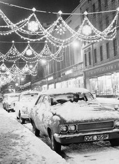 cars parked on the side of a snowy street in front of buildings with christmas lights