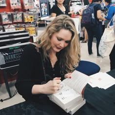 a woman sitting at a table with an open book in front of her and writing on it