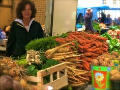 a woman standing in front of a pile of carrots and other vegetables at a market