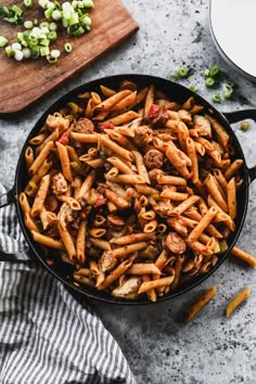 a skillet filled with pasta and meat on top of a table next to a cutting board