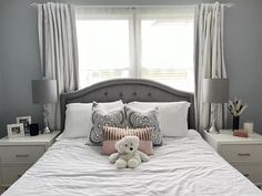 a bed with white sheets, pillows and a teddy bear on it in front of two windows