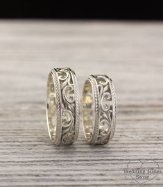 two wedding bands with intricate designs on them sitting on top of a wooden table next to each other