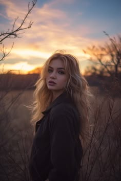 a woman standing in tall grass with the sun setting behind her and looking off into the distance