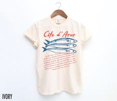 Set sail for style with this vintage-inspired Côte d'Azur graphic tee! Perfect for seafood lovers and travel enthusiasts, it captures the coastal charm and vibrant flavors of the French Riviera. The text on the bottom reads, "The Côte d'Azur, or French Riviera, is a glamorous Mediterranean destination known for its stunning beaches, luxurious resorts, and charming coastal towns. In addition to its breathtaking beauty, the region is famous for its fresh seafood, particularly sardines, sea bass, a French Beach Style, Italian Graphics, Vintage Aesthetic Retro, French Beach, Travel Tees, Aesthetic Retro, Retro Graphic Tees, Coastal Charm, Sea Bass