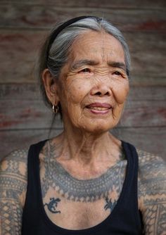 Portrait d'une femme âgée asiatique souriante aux cheveux gris, portant un débardeur noir, avec des tatouages tribaux traditionnels ornant son cou et sa poitrine, photographiée devant un mur texturé Oldest Tattoo, Whang Od, Native Women, Filipino Tattoos, Tattoo Techniques, Indigenous Women, Old Tattoos, Aesthetic Tattoo, Old Woman