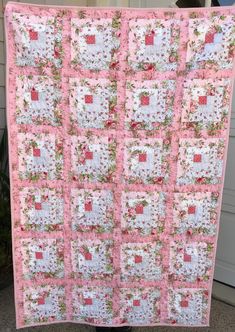 a pink and white quilt hanging from the side of a house with flowers on it