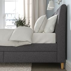 a bed with pillows and blankets on it in front of a window next to a potted plant