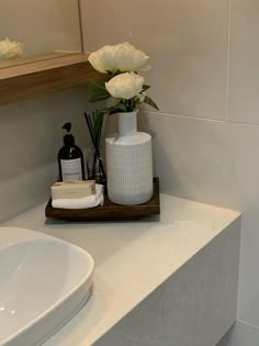 a white vase with flowers and soap on a wooden tray next to a bathroom sink