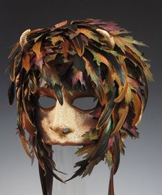 a mask with feathers on it is shown in front of a gray background and the face has been made out of leaves